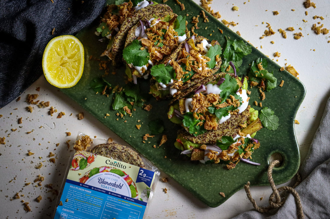 Tacos mit Caulito veganen Toasties. Caulito Vegane Tacos werden auf einem rustikalen grünen Brett mit Zitrone platziert. Eine Packung Blumenkohl-Toasties in einer biologisch abbaubaren Verpackung.