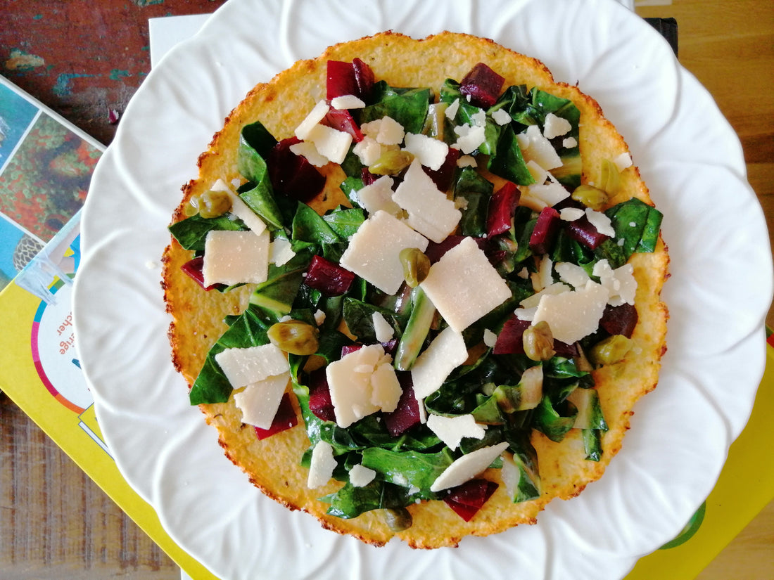 Low Carb glutenfreier Blumenkohl-Pizzaboden mit Spinat, Kapern und Käse auf einem weißen Porzellanteller, geschmackvoll auf einem Holzbrett platziert.